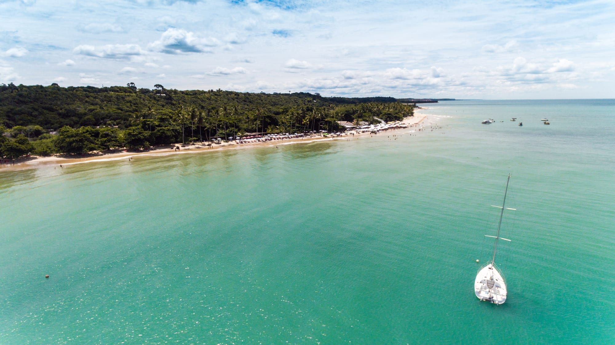 Vista aérea da Praia dos Coqueiros, em Trancoso, com mar verde-azulado, faixa de areia clara, mata nativa e um veleiro ancorado