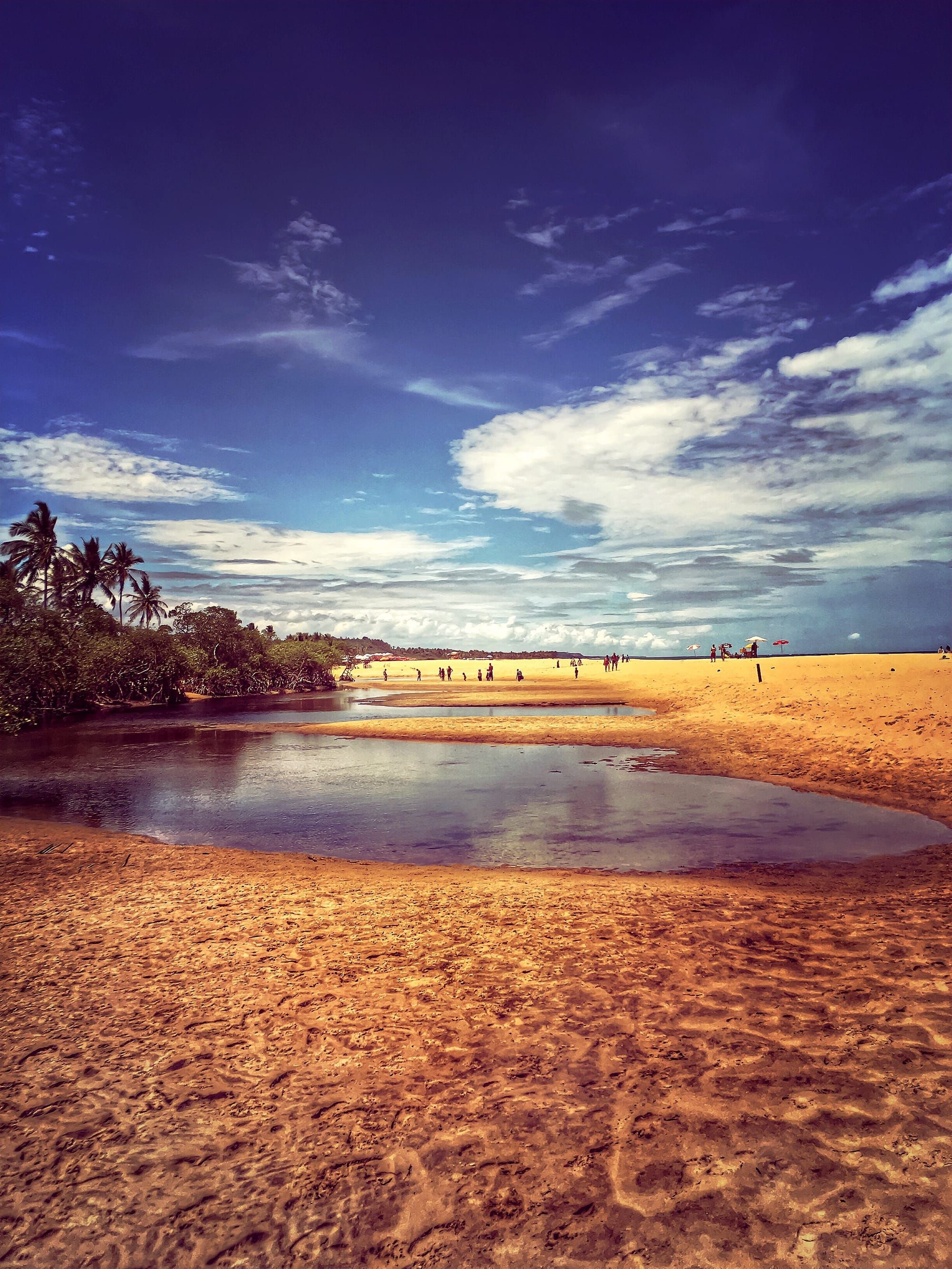 Encontro do rio com o mar em Trancoso, com a água refletindo o céu do fim de tarde e coqueiros ao fundo